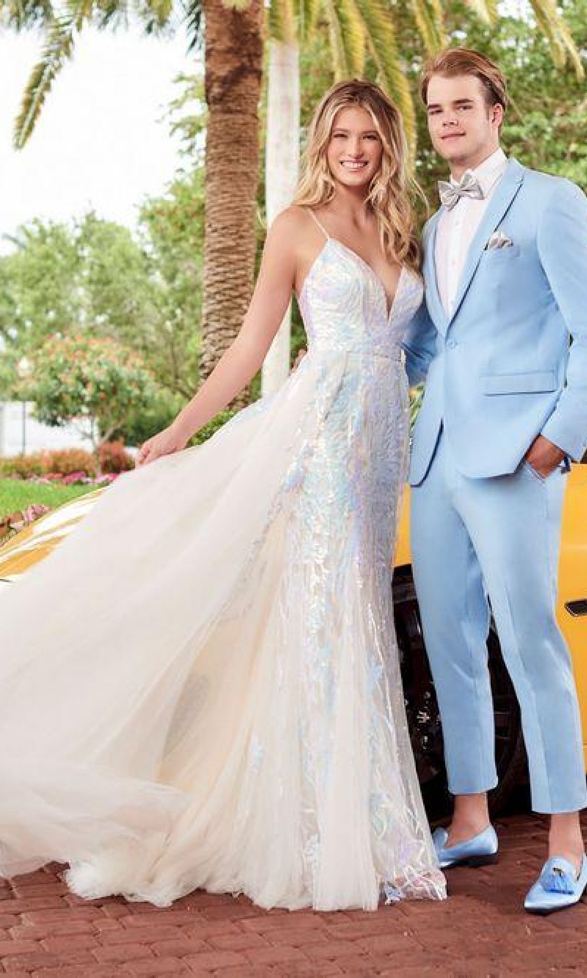 A woman in the Tiffany 16944 Long Sequin Prom Dress stands beside a man in a light blue suit with a bow tie; together they pose outdoors near a yellow car and palm trees.
